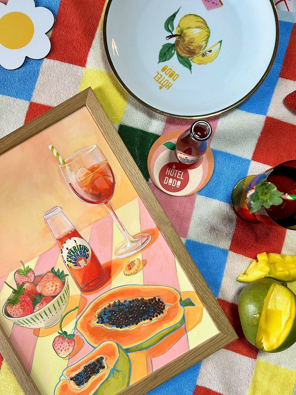 A vibrant Indian summer picnic setup featuring a colorful checkered blanket, lemon-patterned plate, L'Aperitivo Leopardo Giclée Art Print by Hôtel Dodo, vermouth with a straw, fresh fruit, and a "Hôtel Deux" coaster.
