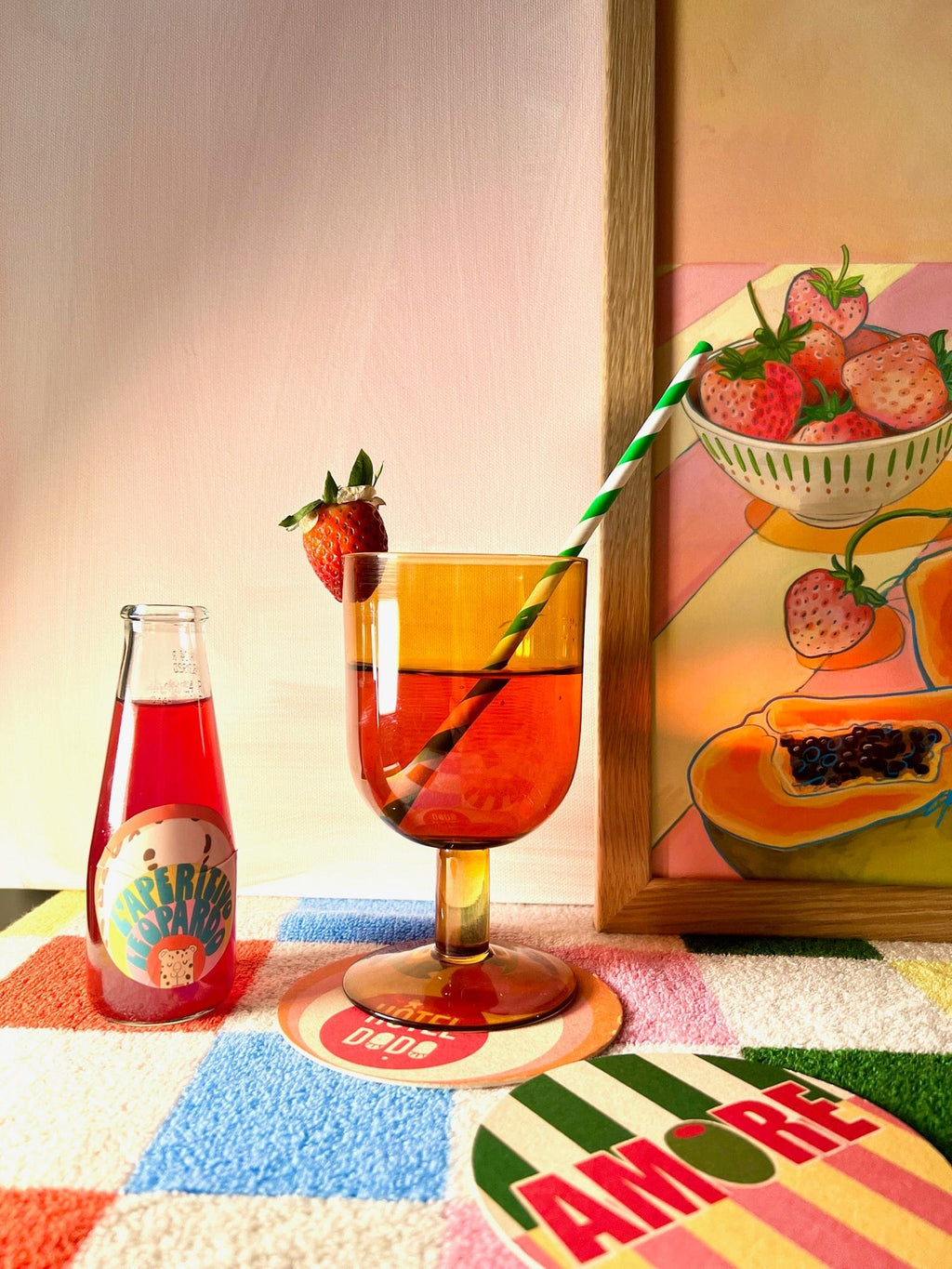 A glass of red vermouth with a strawberry and striped straw, the L'Aperitivo Leopardo Giclée Art Print by Hôtel Dodo, and a "HAPPY PARADISE" bottle create a vibrant setup—ideal for an Indian summer picnic at Hôtel Dodo.