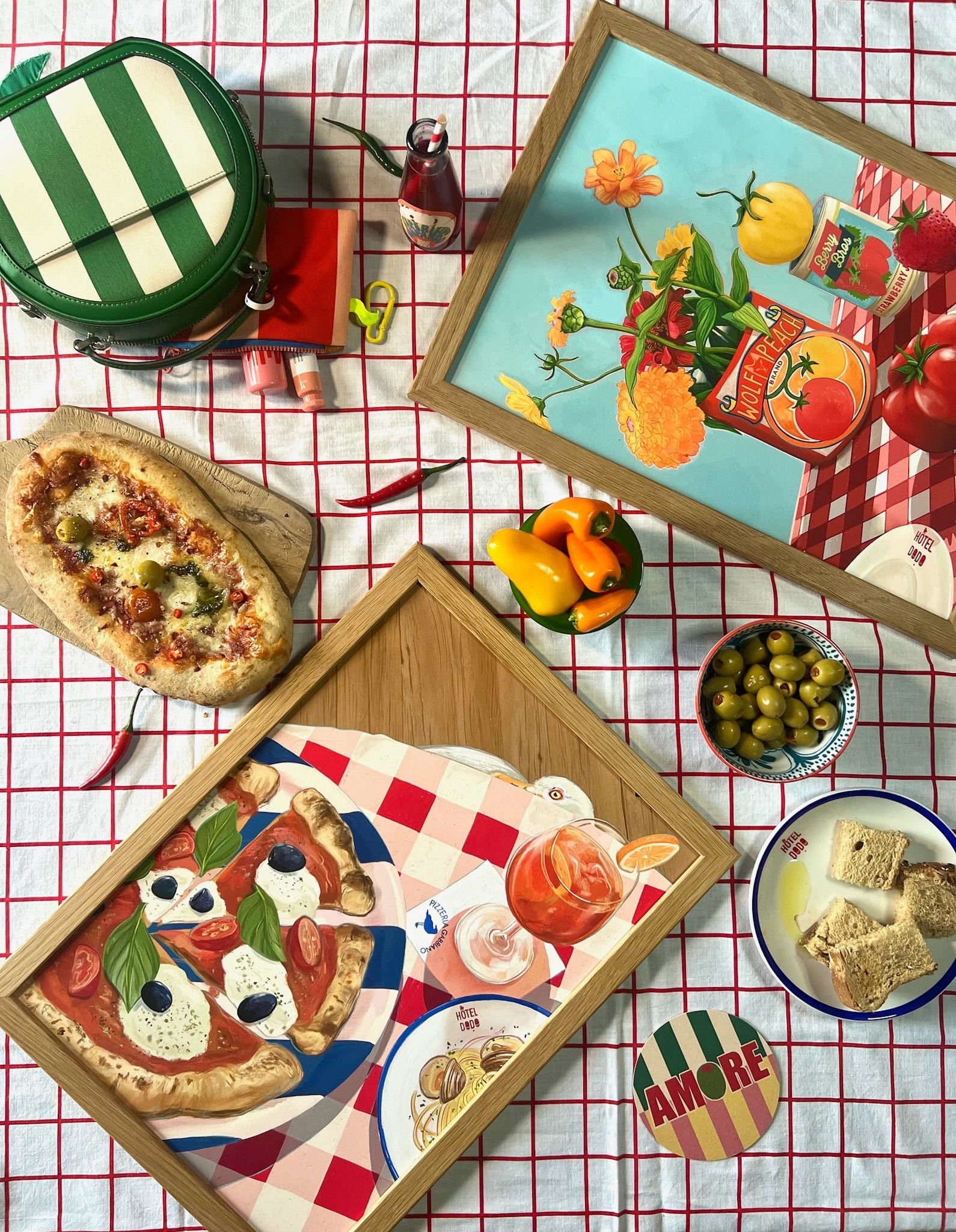 A tabletop displays the Hôtel Dodo "You Say Tomato I Say Wolf Peach" Giclée Art Print with a grid-patterned cloth, pizza artwork topped with tomato slices, and neatly arranged dining items including bread, olives, and a green-striped bag.