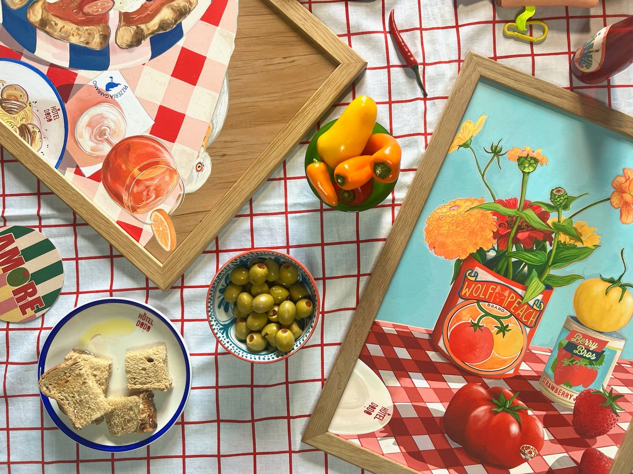 A table with a red and white grid cloth displays Hôtel Dodo's "You Say Tomato I Say Wolf Peach" Giclée Art Print, framed among drinks, olives, bread, yellow peppers, nightshade tomatoes, and a plate reading "AMORE.