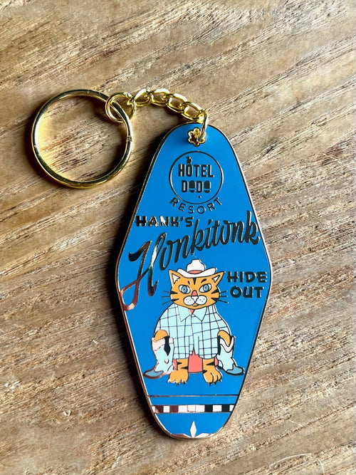 A Hank's Honkytonk Hideout Keyring by Hôtel Dodo, featuring a cat dressed as a cowboy on an enamel tag, sits on wood, evoking rustic log cabin charm.