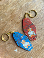 Two Hôtel Dodo "Hank's Honkitonk Hideout" keyrings rest on wood, featuring a vintage motel key design. One red, one blue, each displays a cartoon cat in plaid with messages "Stay Fresh" and "Hide Out," capturing the whimsical charm of a secluded log cabin.
