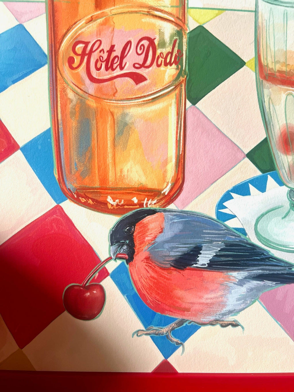 A bullfinch with a cherry stands on a vibrant checkered surface beside a "Hôtel Dodo" bottle in the Fat Finch Soda Giclée Art Print, blending retro summer nostalgia with whimsical charm.