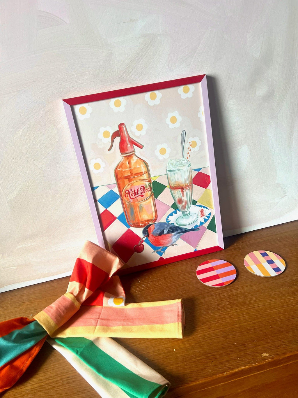 The Fat Finch Soda Giclée Art Print by Hôtel Dodo rests on a wooden surface beside a striped scarf and two round, striped coasters—capturing the cozy charm of Hôtel Dodo.