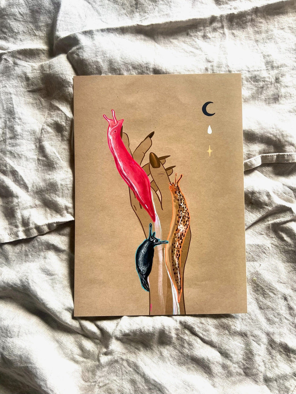 The Midnight Slugs Matte Art Print by Beauty and the Beastie features a hand with painted nails holding three colorful slugs under celestial symbols, all set on brown paper atop wrinkled fabric.