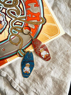 Two vintage-style keychains, crafted by Hôtel Dodo, rest on fabric. One blue and one red, they each display a cartoon cat with text. Nearby, Hank's Honkitonk Hideout Keyring reveals a colorful circular design inspired by an enamel tag from a cozy log cabin.