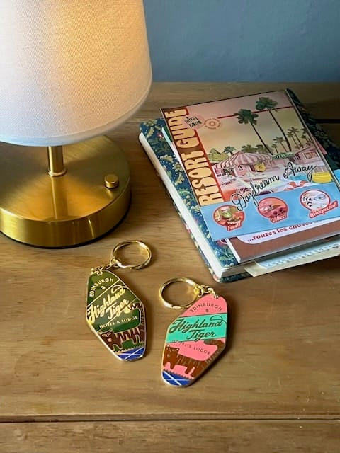 A lamp, a travel guide, and two colorful Highland Tiger Hotel Keyring Dark by Essential Edinburgh rest on a wooden surface—perfect souvenirs from the Highland Tiger Hotel.