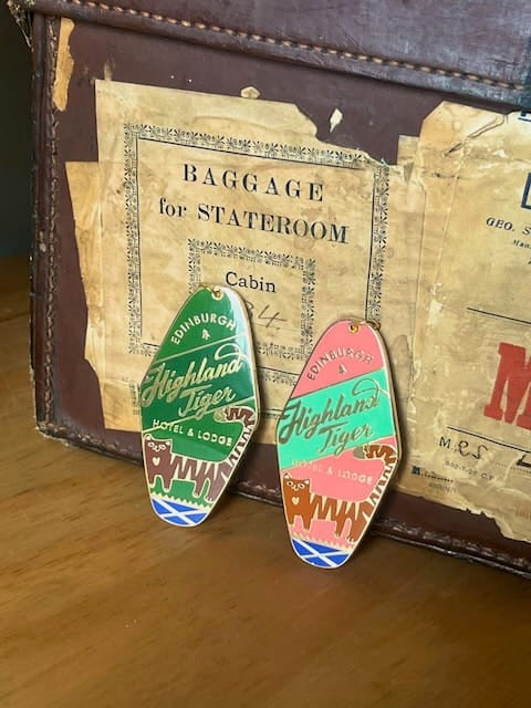 Two colorful Highland Tiger Hotel Keyring Dark keychains by Essential Edinburgh rest against a vintage suitcase with worn travel labels, adding a charming bag charm touch to any traveler’s collection.