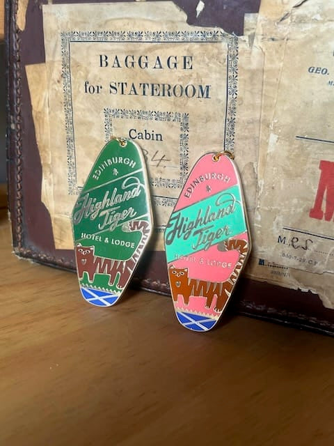 Two “Highland Tiger Hotel Keyring Dark” enamel keyrings by Essential Edinburgh are displayed in front of a vintage suitcase with a worn “Baggage for Stateroom” label, each enamel tag adding a touch of charm reminiscent of classic hotel bag charms.