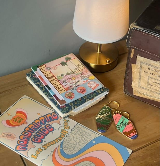 A wooden desk displays a lamp, a vintage suitcase, two keychains—including the Essential Edinburgh Highland Tiger Hotel Keyring Dark—notebooks, and a colorful “Roadtripping Guide” leaflet with a retro design from the Highland Tiger Hotel.