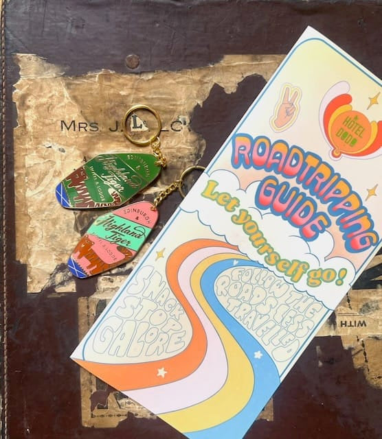 A brochure titled “Roadtripping Guide” and two motel keychain keyrings—including the Essential Edinburgh Highland Tiger Hotel Keyring Dark—rest on a worn book cover labeled “Mrs. J.L.C.”.
