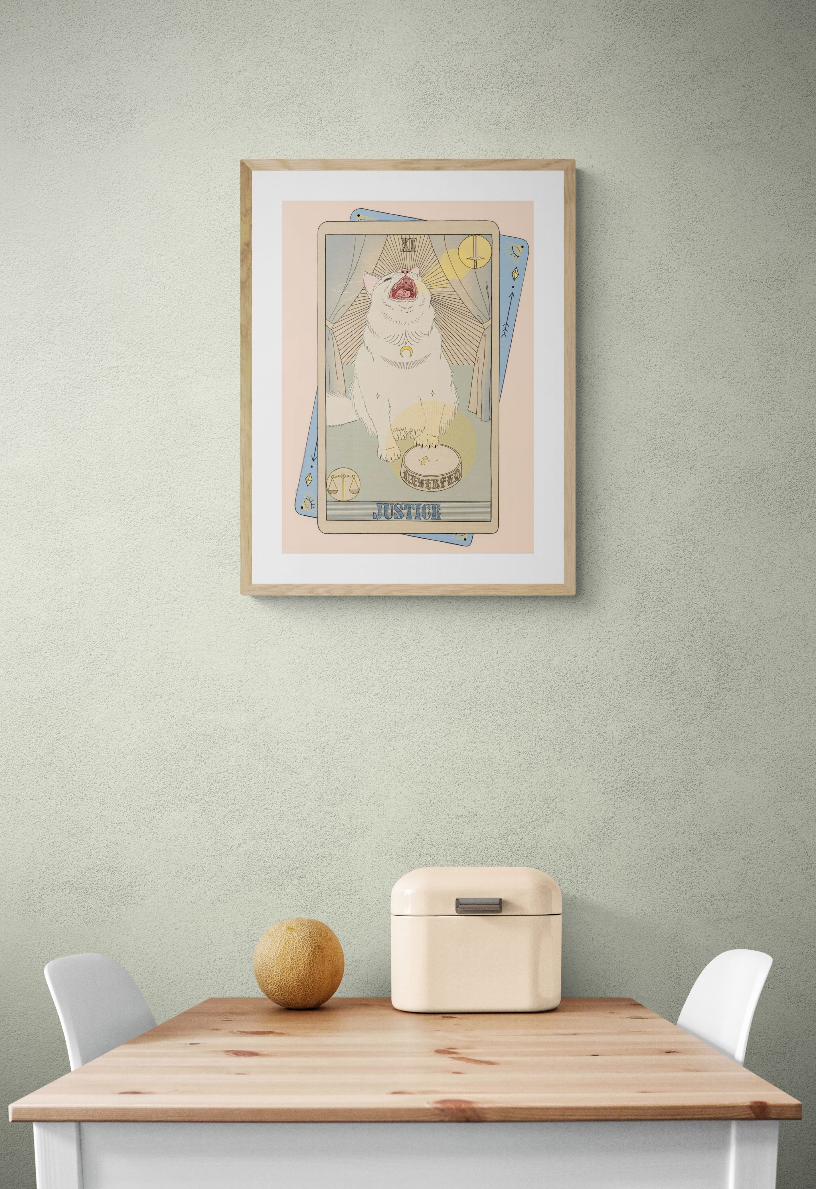 The Justice Matte Art Print by Tarot Cats—a collectible featuring a white cat in vintage tarot card style—hangs on a light green wall above a small table, adding charm and whimsy to your space.