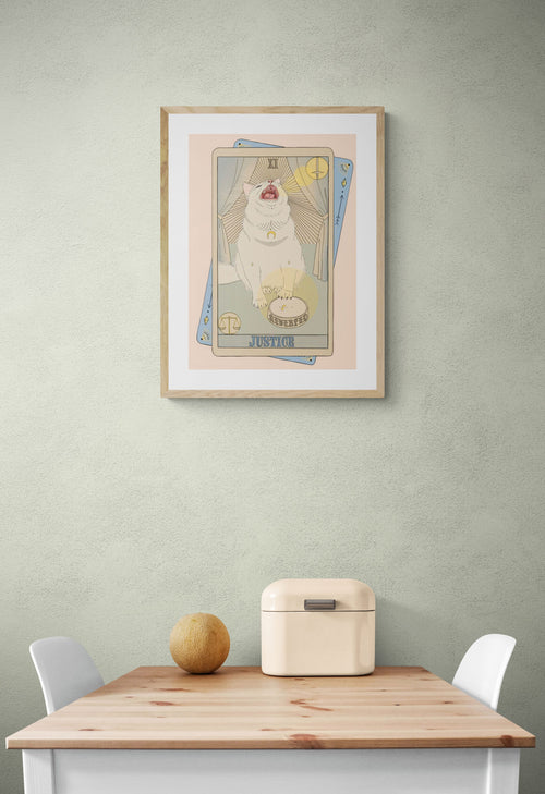 The Justice Matte Art Print by Tarot Cats—a collectible featuring a white cat in vintage tarot card style—hangs on a light green wall above a small table, adding charm and whimsy to your space.