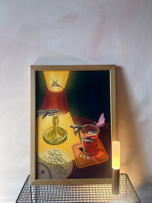 The Hôtel Dodo Lamp Lounge Giclée Art Print, featuring a lit lamp, vermouth glass, moths, and coasters, is displayed on a wire shelf with a small cylindrical light in front against a pale wall.