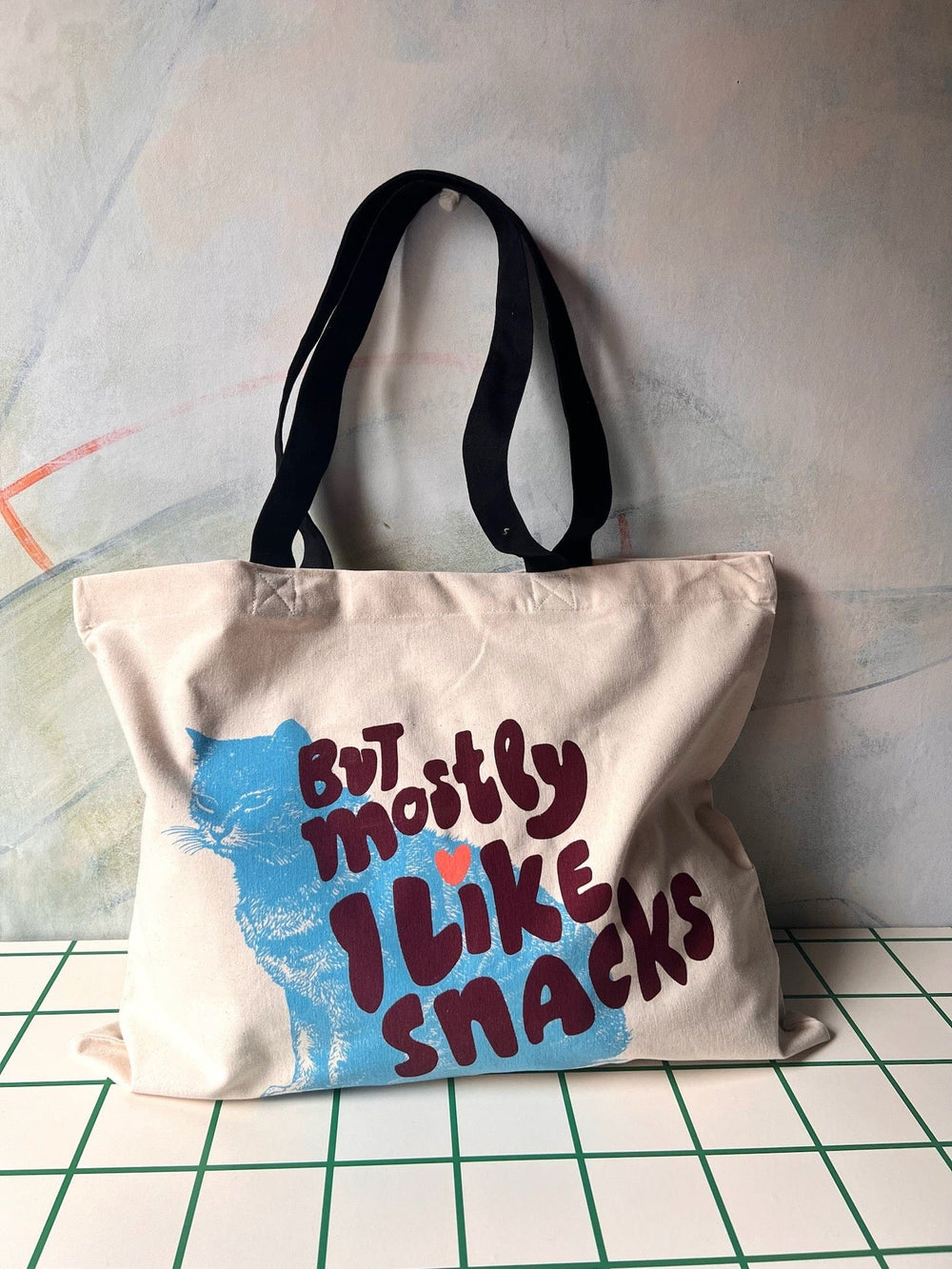 The "But Mostly I Like Cats" Shopping & Beach Bag by Bags by diedododa features a cotton canvas design with black handles, a blue cat graphic, and the bold red phrase "But mostly I like snacks," making it an ideal tote for snack lovers.