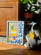 The Amalfi Limoni Giclée Art Print by Hôtel Dodo rests framed on a wooden floor beside a gray bag with a lemon keychain and a potted plant, capturing the sunny charm of the Amalfi coast.