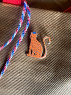 A charming ginger Léopard Roux Pin by Pins by diedododa features a freckled face and "Leopard Roux has Bonnet" on its body, attached with a stylish red and blue striped rope.