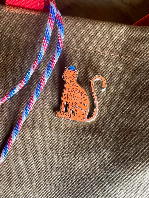 A charming ginger Léopard Roux Pin by Pins by diedododa features a freckled face and "Leopard Roux has Bonnet" on its body, attached with a stylish red and blue striped rope.