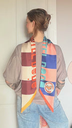 A person with short hair, wearing a taupe t-shirt and light blue jeans, faces away from the camera as the colorful Hotel Dodo Scarf by diedododa, featuring bold patterns, drapes lightly over their shoulders.