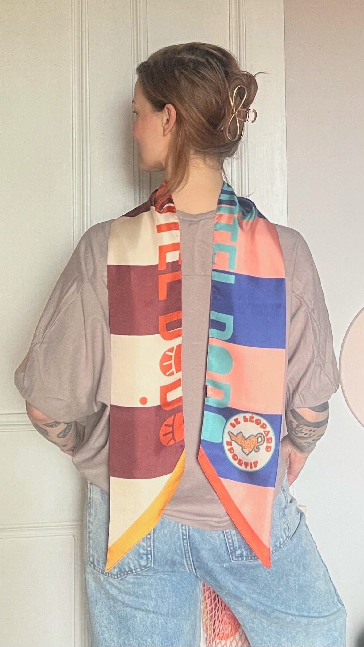 A person with short hair, wearing a taupe t-shirt and light blue jeans, faces away from the camera as the colorful Hotel Dodo Scarf by diedododa, featuring bold patterns, drapes lightly over their shoulders.