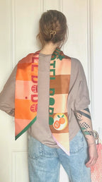 A person with light brown hair in a clip, wearing a beige top and light blue jeans, stands facing away; the diedododa Hotel Dodo Scarf, featuring colorful geometric and fruit designs, is draped over their shoulders.