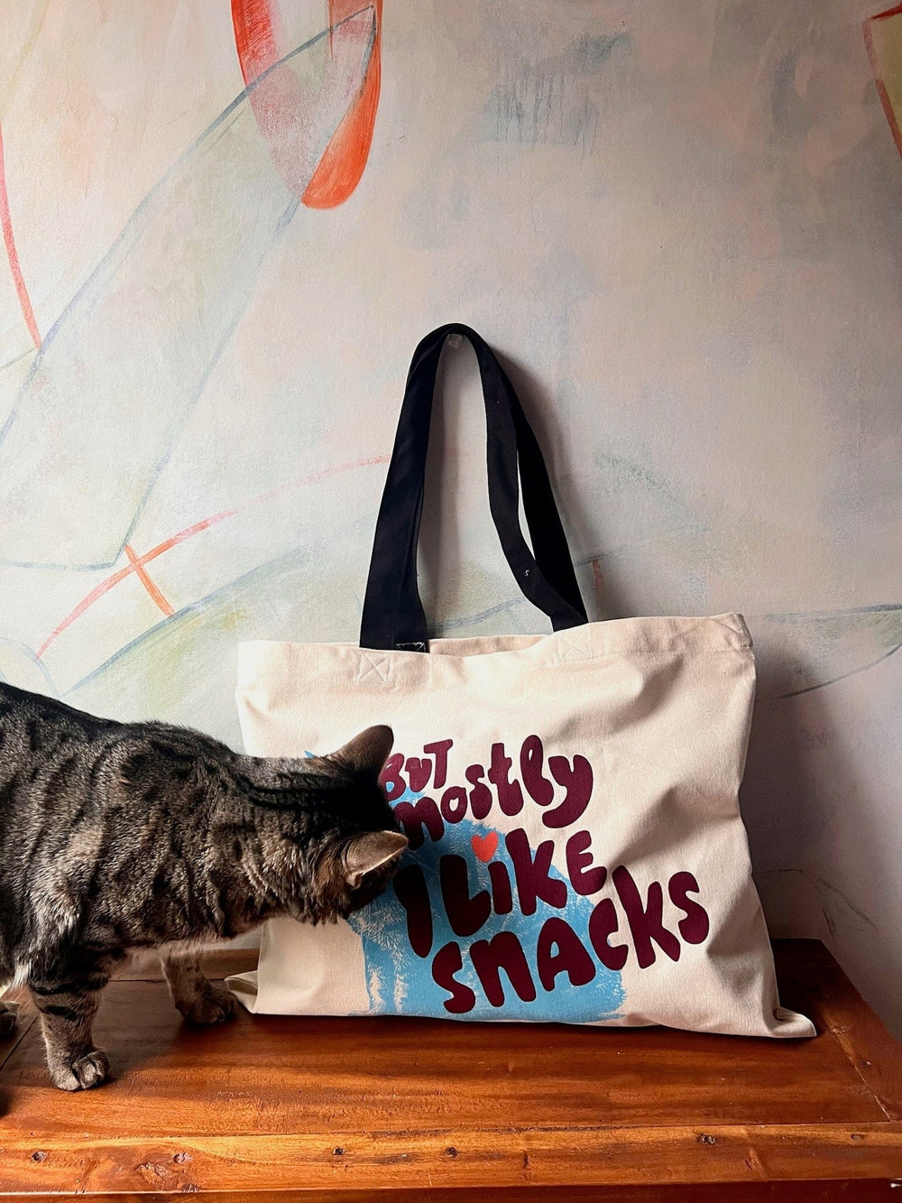 A striped cat sniffs a cotton canvas bag from Bags by diedododa, named the "But Mostly I Like Cats Shopping & Beach Bag." The tote rests on a wooden surface against a painted wall.