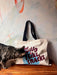 A striped cat sniffs a cotton canvas bag from Bags by diedododa, named the "But Mostly I Like Cats Shopping & Beach Bag." The tote rests on a wooden surface against a painted wall.