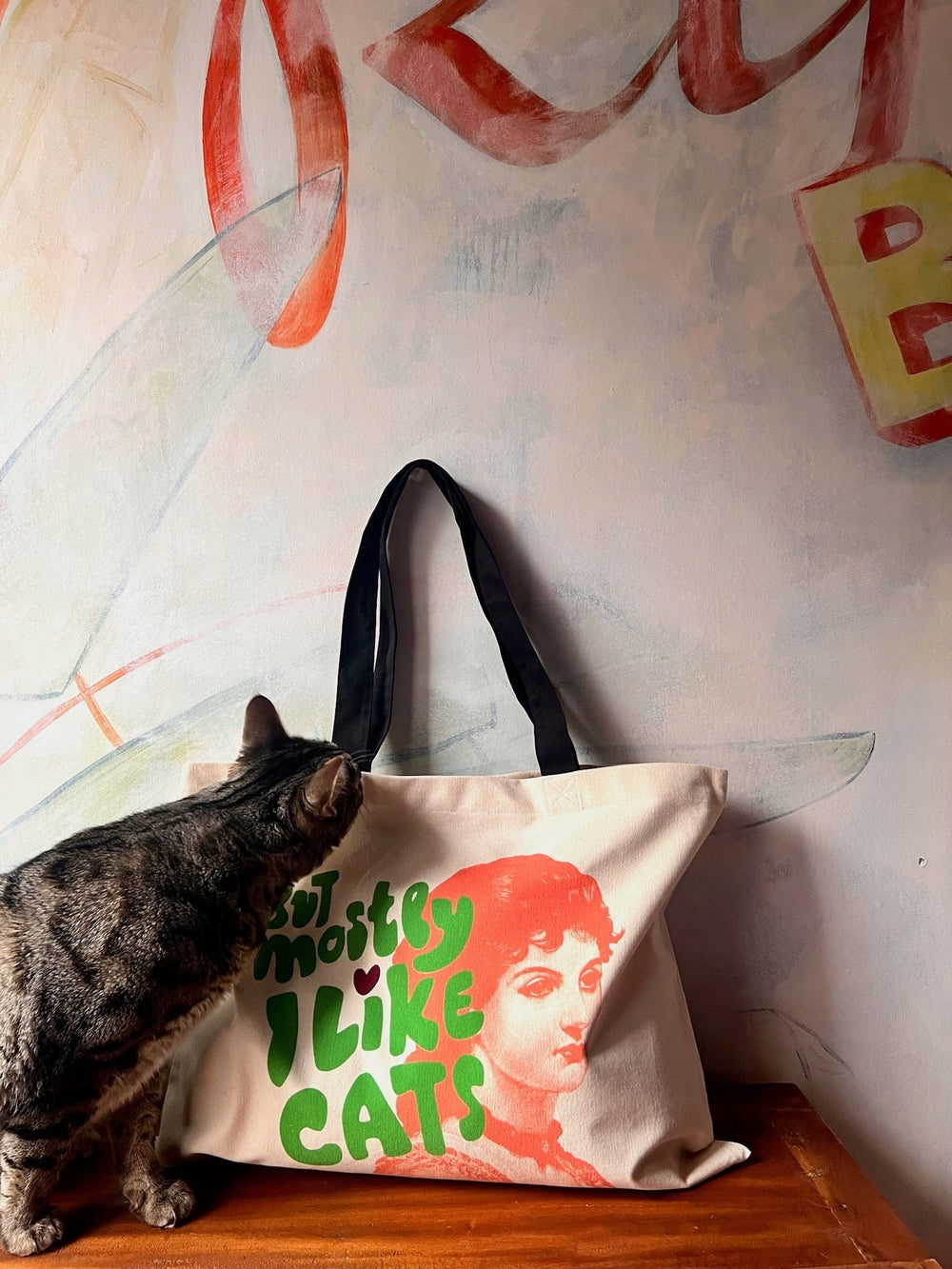 A curious cat sniffs a cotton canvas "But Mostly I Like Cats Shopping & Beach Bag" by Bags by diedododa, set against a colorful wall.