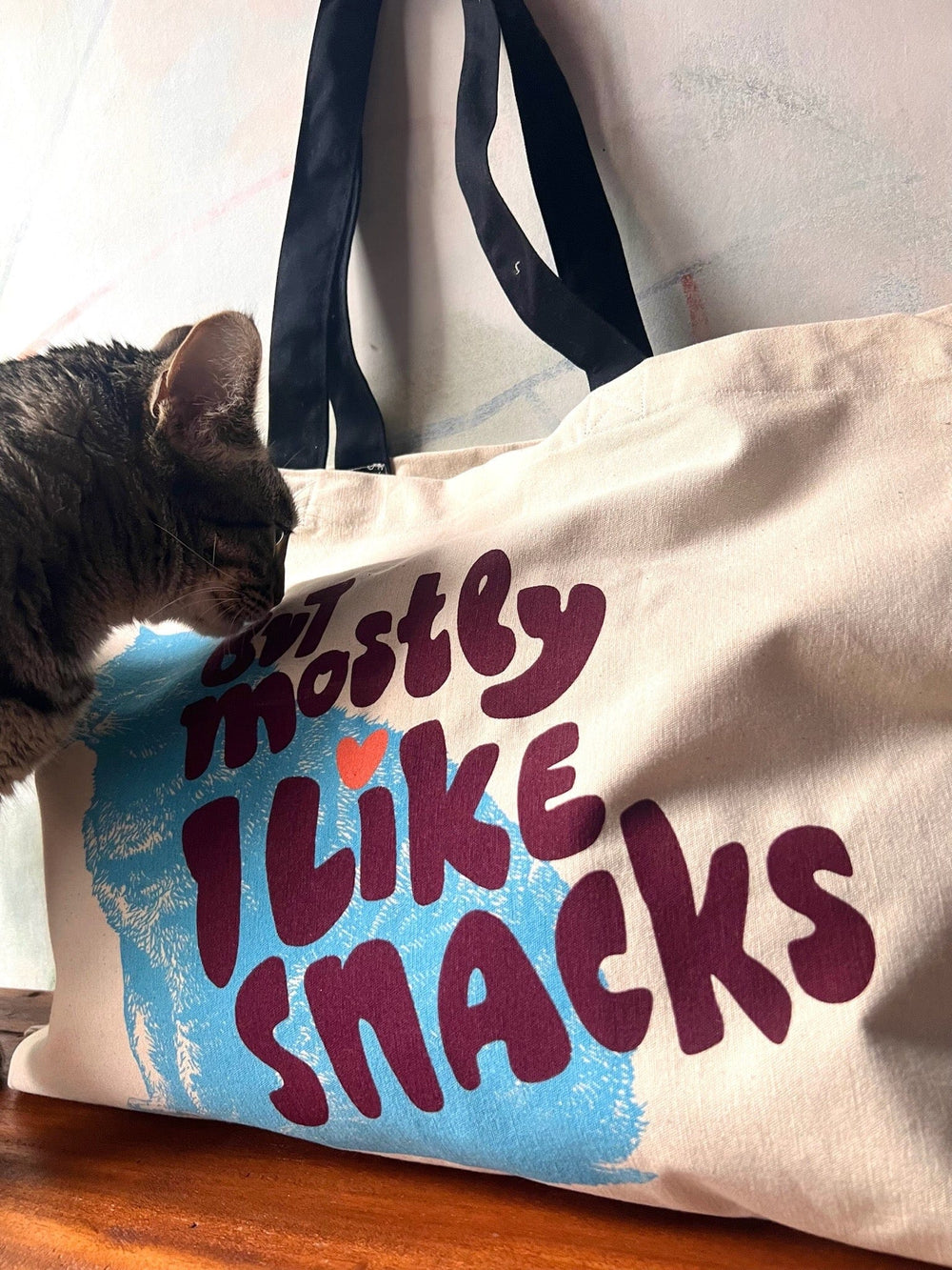 A curious cat sniffs a cotton canvas bag adorned with black handles. The tote, created by Bags by diedododa, reads "But Mostly I Like Cats" in bold, colorful letters.