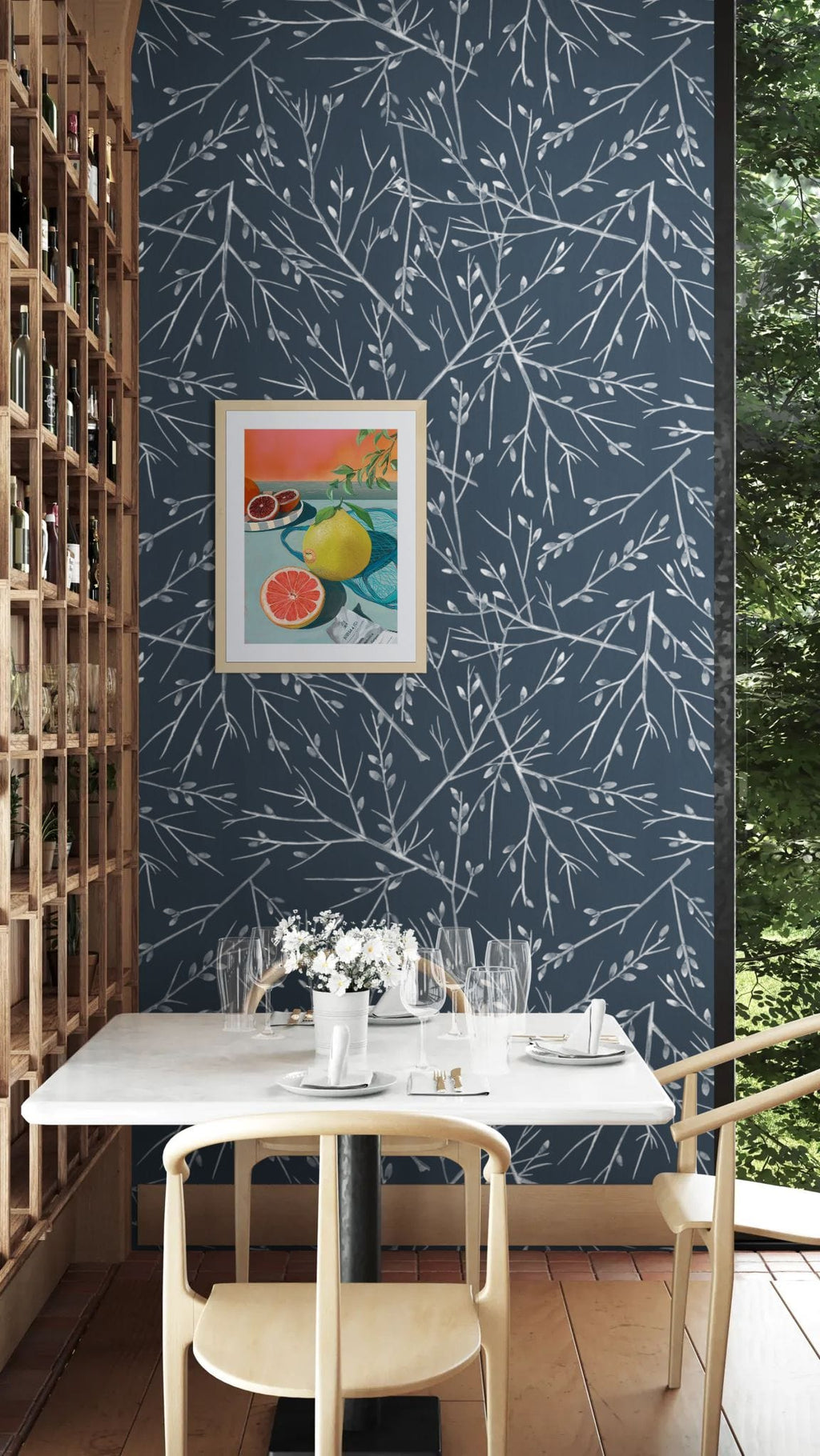 A small dining table with curated settings and two chairs sits by a wall clad in dark blue, white-branch wallpaper and features Hôtel Dodo's Mellow Pomelo Giclée Art Print; a wine rack is on the left, and the window shows greenery outside.