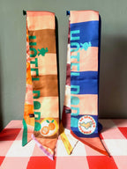 Two vibrant, patterned Hotel Dodo Scarves by diedododa, crafted from 100% silk with "HOTEL BOB" text and fruit-themed patches, are displayed upright on a pink checkered surface against a gray background.