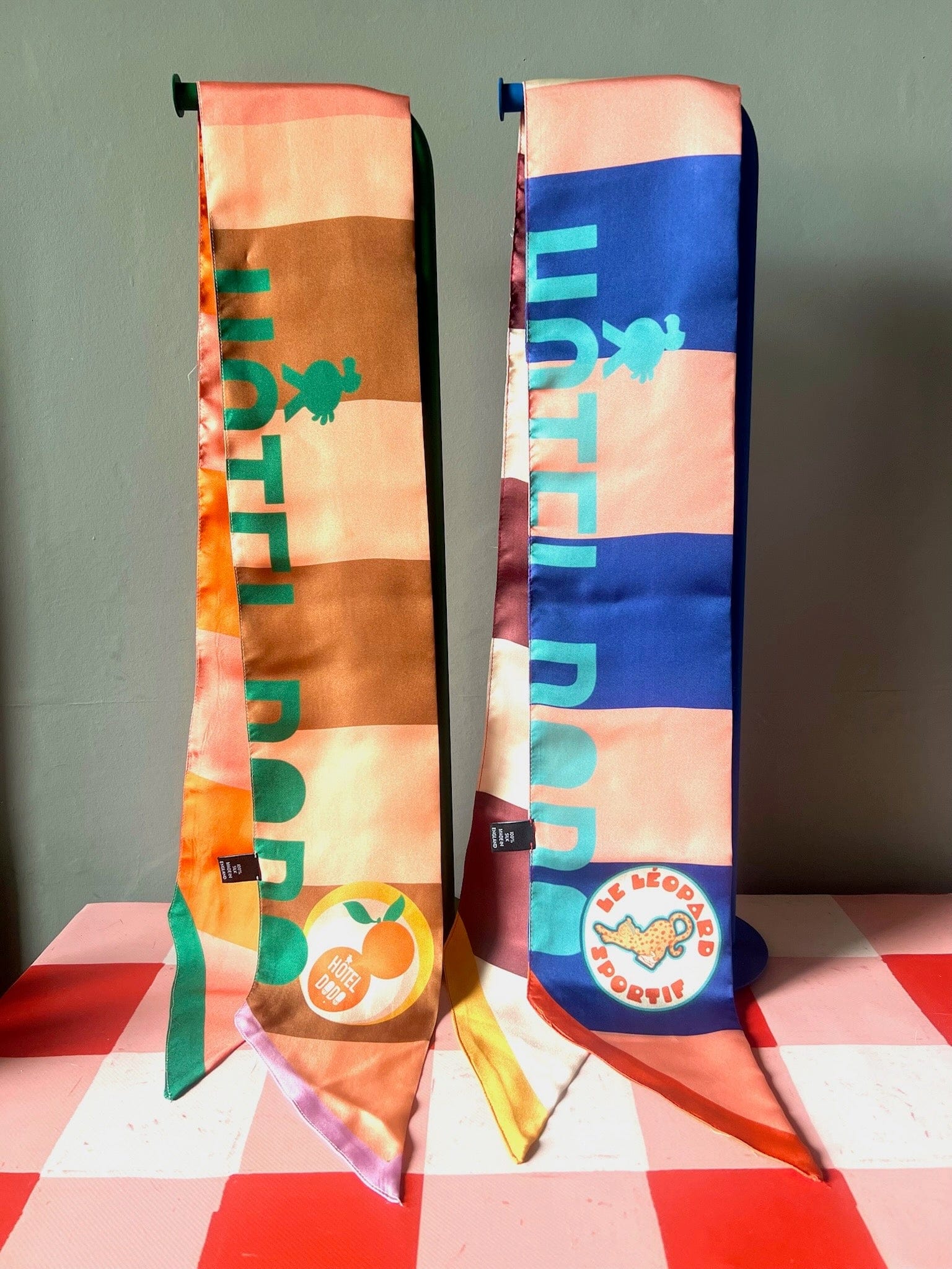 Two vibrant, patterned Hotel Dodo Scarves by diedododa, crafted from 100% silk with "HOTEL BOB" text and fruit-themed patches, are displayed upright on a pink checkered surface against a gray background.