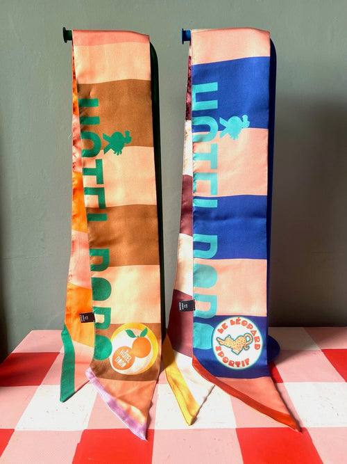 Two vibrant, patterned Hotel Dodo Scarves by diedododa, crafted from 100% silk with "HOTEL BOB" text and fruit-themed patches, are displayed upright on a pink checkered surface against a gray background.