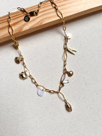 The Seaside Treasures Necklace from Necklaces features a gold chain adorned with charming seashells, a starfish, and delicate leaves, beautifully displayed against a light fabric background framed in wood. This exquisite 18K gold-plated piece is ideal for seafood enthusiasts wanting to infuse their jewelry collection with a coastal vibe.