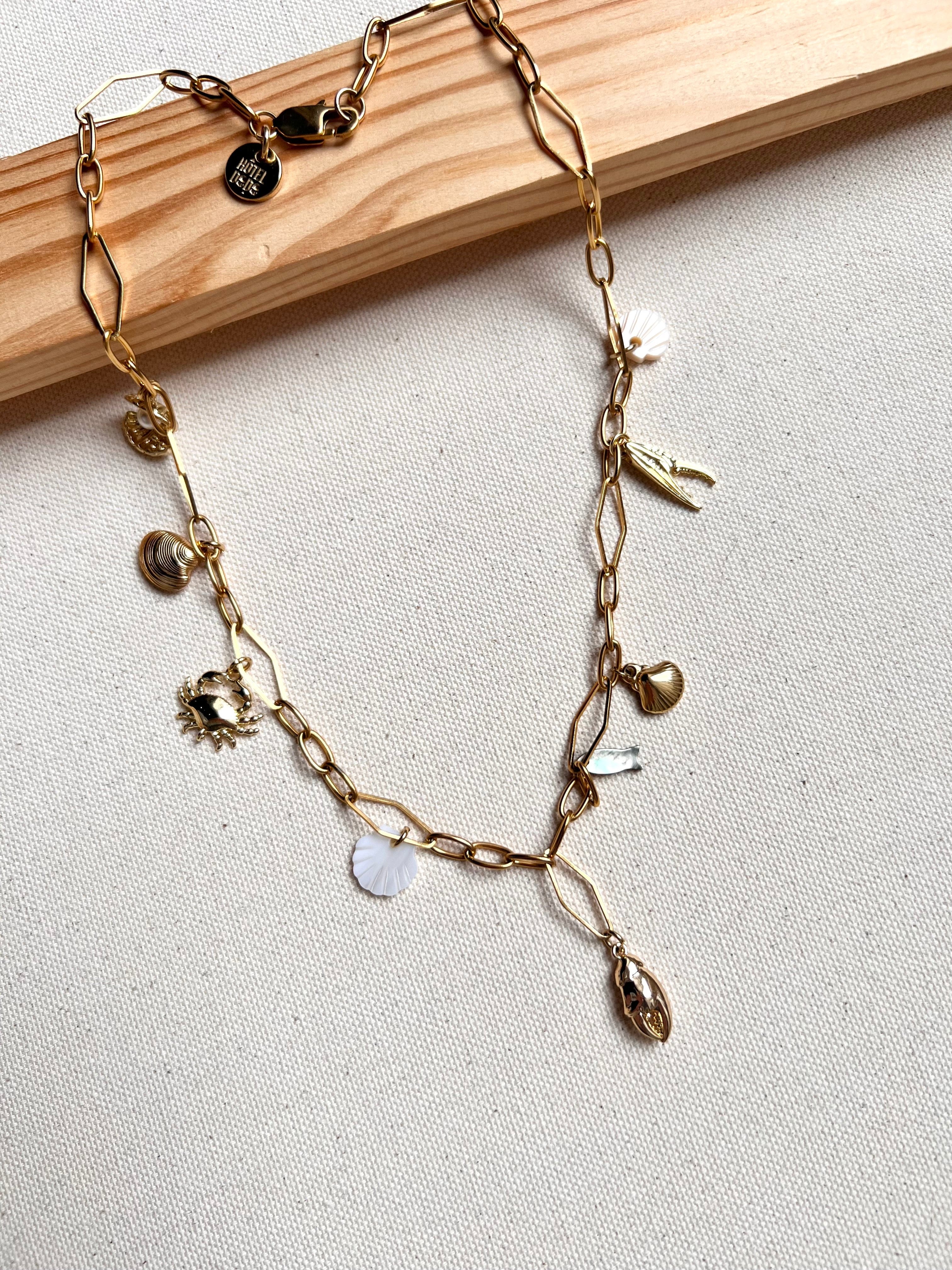 The Seaside Treasures Necklace from Necklaces features a gold chain adorned with charming seashells, a starfish, and delicate leaves, beautifully displayed against a light fabric background framed in wood. This exquisite 18K gold-plated piece is ideal for seafood enthusiasts wanting to infuse their jewelry collection with a coastal vibe.