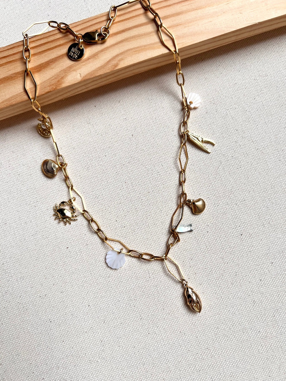 The Seaside Treasures Necklace from Necklaces features a gold chain adorned with charming seashells, a starfish, and delicate leaves, beautifully displayed against a light fabric background framed in wood. This exquisite 18K gold-plated piece is ideal for seafood enthusiasts wanting to infuse their jewelry collection with a coastal vibe.