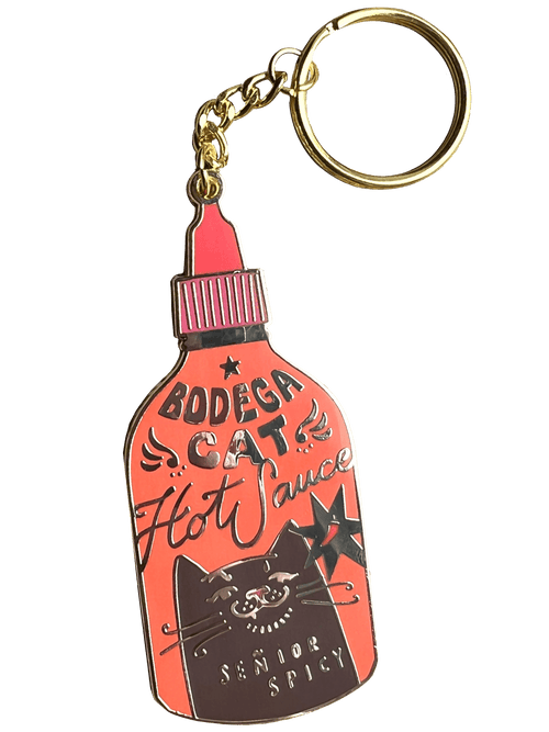 The Hôtel Dodo Bodega Cat Hot Sauce Keyring is a keychain and bag charm shaped like a hot sauce bottle, featuring a cartoon bodega cat with “Bodega Cat Hot Sauce” and “Senior Spicy” on an enamel tag.