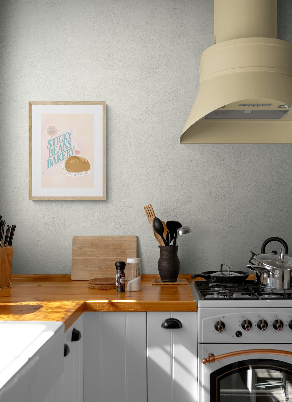 A kitchen with a wooden countertop, utensils in a jar, cutting boards, spice jar, gas stove with a kettle, and the Sticky Beans Bakery Loaf Of Your Life Matte Art Print by International Pestsellers framed on a light-colored wall.