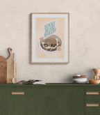 The Sticky Beans Bakery Loaf In A Hopeless Place Matte Art Print by International Pestsellers hangs above a green kitchen counter with cutting boards, an open book, dishes, and fresh bread, creating an inviting space to relax.