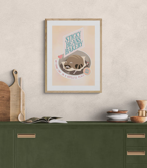 The Sticky Beans Bakery Loaf In A Hopeless Place Matte Art Print by International Pestsellers hangs above a green kitchen counter with cutting boards, an open book, dishes, and fresh bread, creating an inviting space to relax.