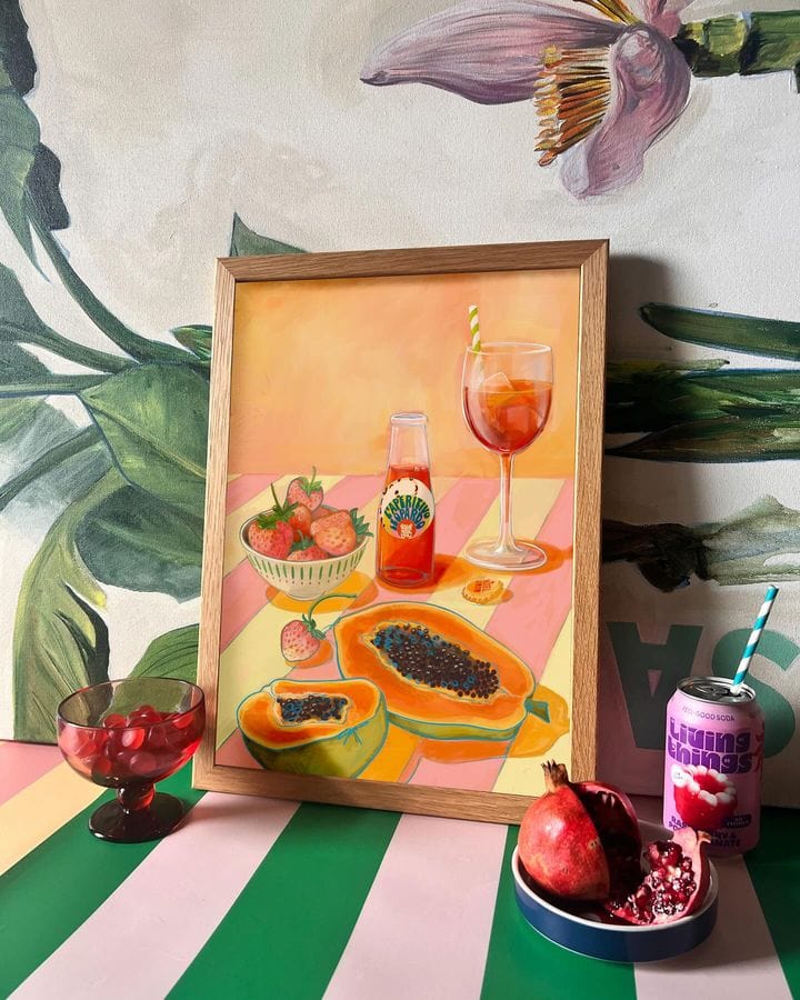 A vibrant still-life setup featuring the L'Aperitivo Leopardo Giclée Art Print by Hôtel Dodo, surrounded by fresh strawberries, papayas, cherries, a pomegranate, and a can of sparkling vermouth on a striped table. This scene evokes the charm of an Indian summer picnic at Hôtel Dodo.