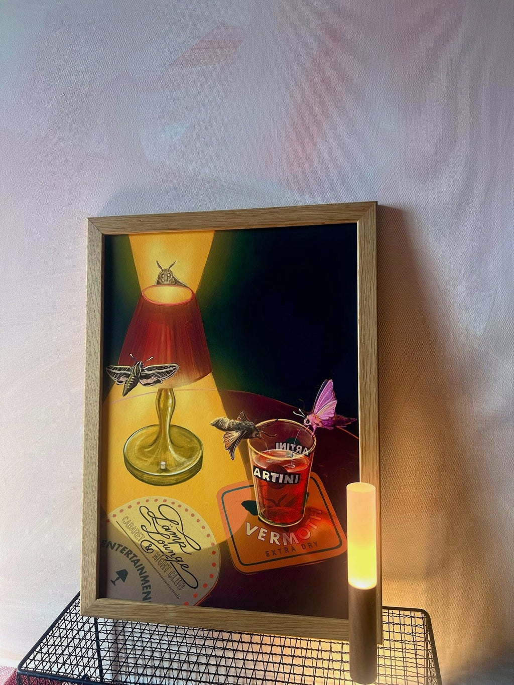 Lamp Lounge Giclée Art Print by Hôtel Dodo features a green lamp, lit lamp, moths, red Artini Vermoth bottle, coaster, and a glowing lamp against a pale pink wall—capturing the mellow ambiance of Lämp Lounge.