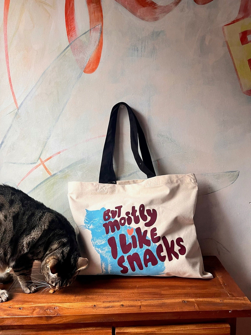 A curious cat sniffs near a shopping and beach tote from Bags by diedododa, featuring the playful phrase "But Mostly I Like Cats." The scene is complemented by a vibrant abstract mural, adding a splash of color to this cozy corner.