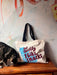 A curious cat sniffs near a shopping and beach tote from Bags by diedododa, featuring the playful phrase "But Mostly I Like Cats." The scene is complemented by a vibrant abstract mural, adding a splash of color to this cozy corner.