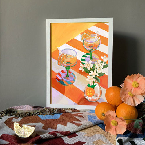 The A L'Orange Giclée Art Print by Hôtel Dodo features two cocktail glasses, white flowers, and an orange on a patterned cloth beside fresh oranges, a lemon wedge, and neroli blossoms—ideal for elevating your table decor.