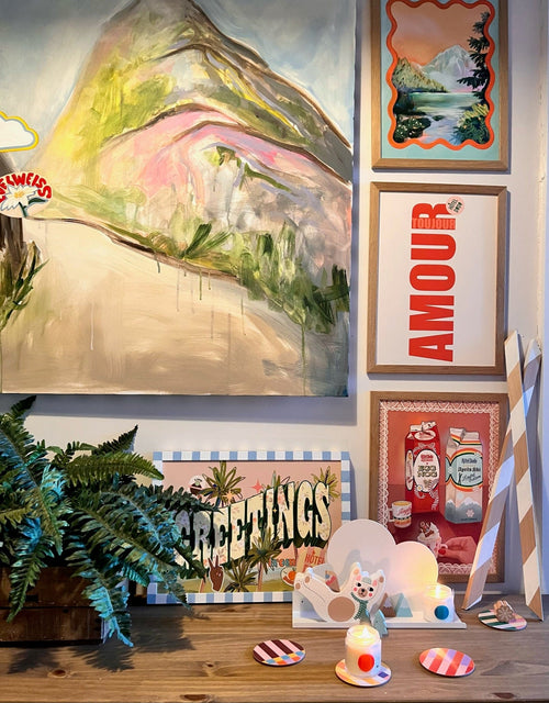 A wooden table holds decorative items, candles, and a plant. On the gallery wall above are colorful artworks and framed prints, including a mountain painting and Shromps' Amour Matte Art Print featuring bold typography.