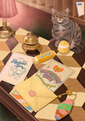 A tabby cat sits on a checkered desk surrounded by stationery, a quirky keychain, a yellow envelope, a candy bar, a cupcake, helpful travel guides titled “Road Tripping Guide” and “Edinburgh Trip,” and the Highland Tiger Lodge Giclée Art Print from Hôtel Dodo.