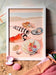 The Tiny Treasures Giclée Art Print by Hôtel Dodo captures a collection of seashells and beach items resting on sand, including a feather, a shell with a pearl, a lobster claw, and dessert on a shell. This artwork invites you to embark on mind travel as if you're lounging at Hôtel Dodo, with shimmers of sea glass adding a touch of oceanic charm.