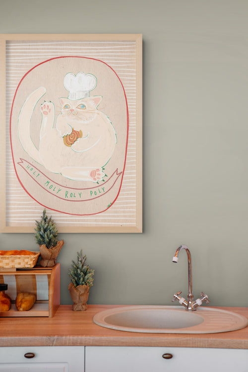 A Roly Poly Matte Art Print from the Sketchy Strays collection, featuring a playful chef cat savoring a jam roly-poly, is framed and hangs above the kitchen sink; on the countertop beneath, fresh bread and small potted trees are arranged.