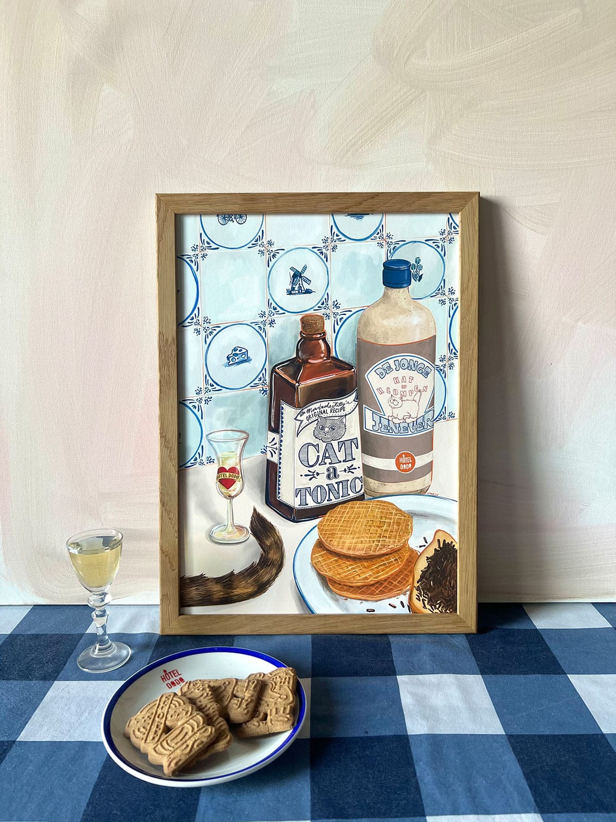 The Dutch Courage Giclée Art Print by Hôtel Dodo depicts vintage bottles, Dutch gin, stroopwafels, and cookies on a blue checkered table—capturing the charm of jenever and classic treats.